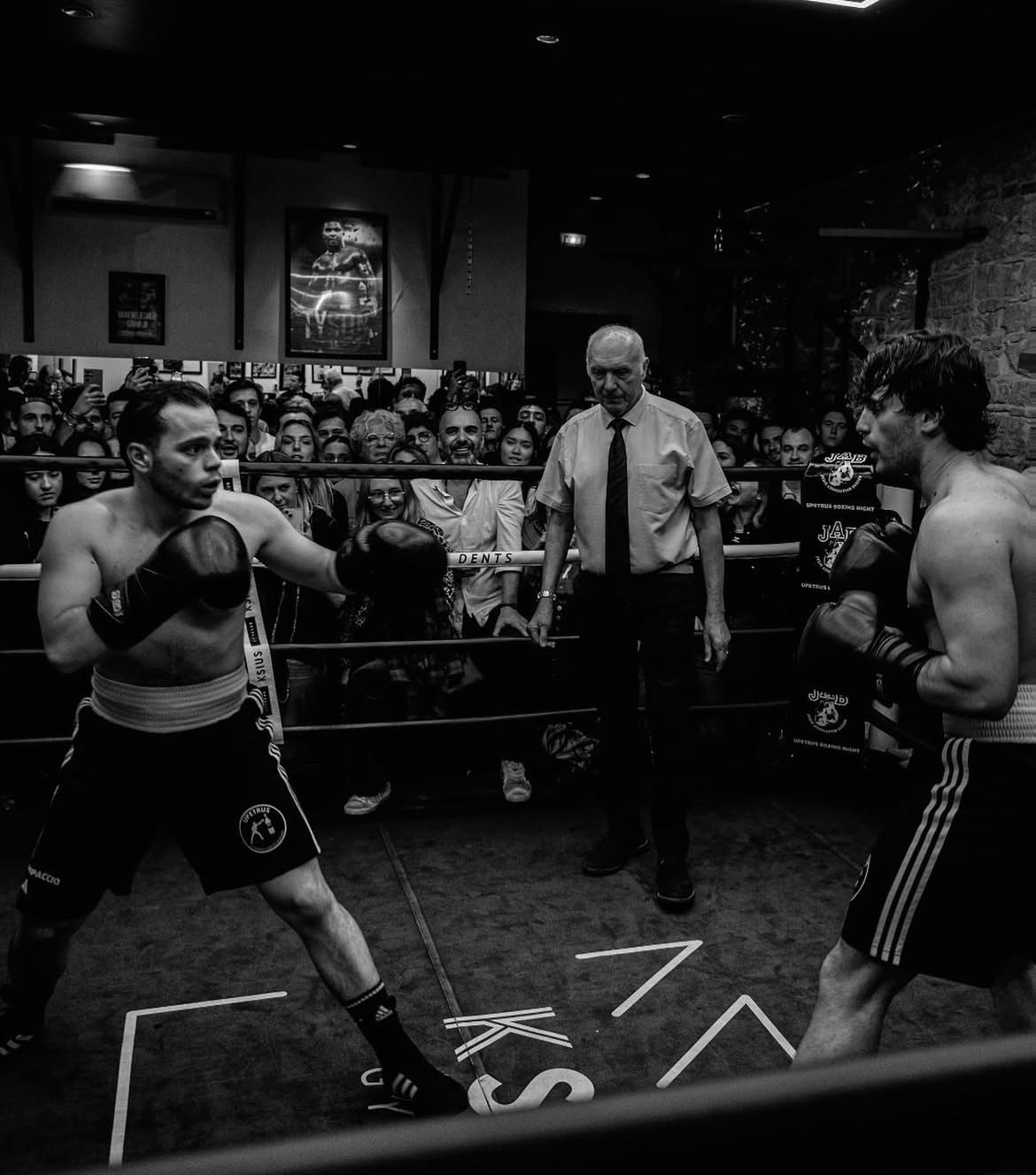 Hugo Rossi sur un ring de boxe, face à son adversaire, l'arbitre entre eux et le public derrière les cordes